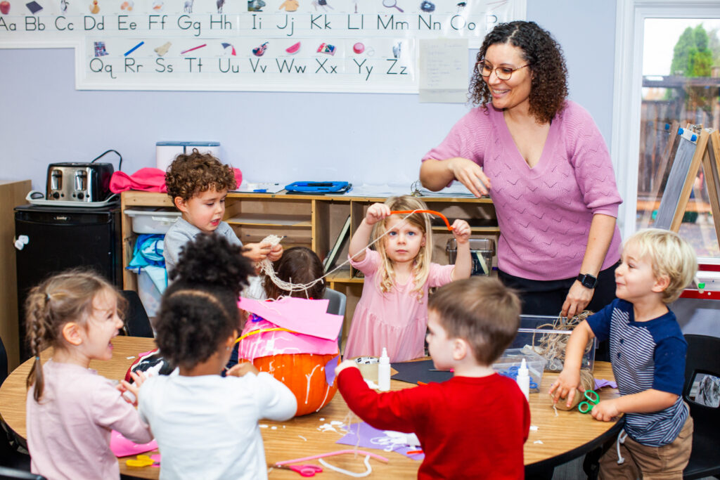 Children creating an art project with a pumpink, scissors, paper, string and glue with a teacher supervising them