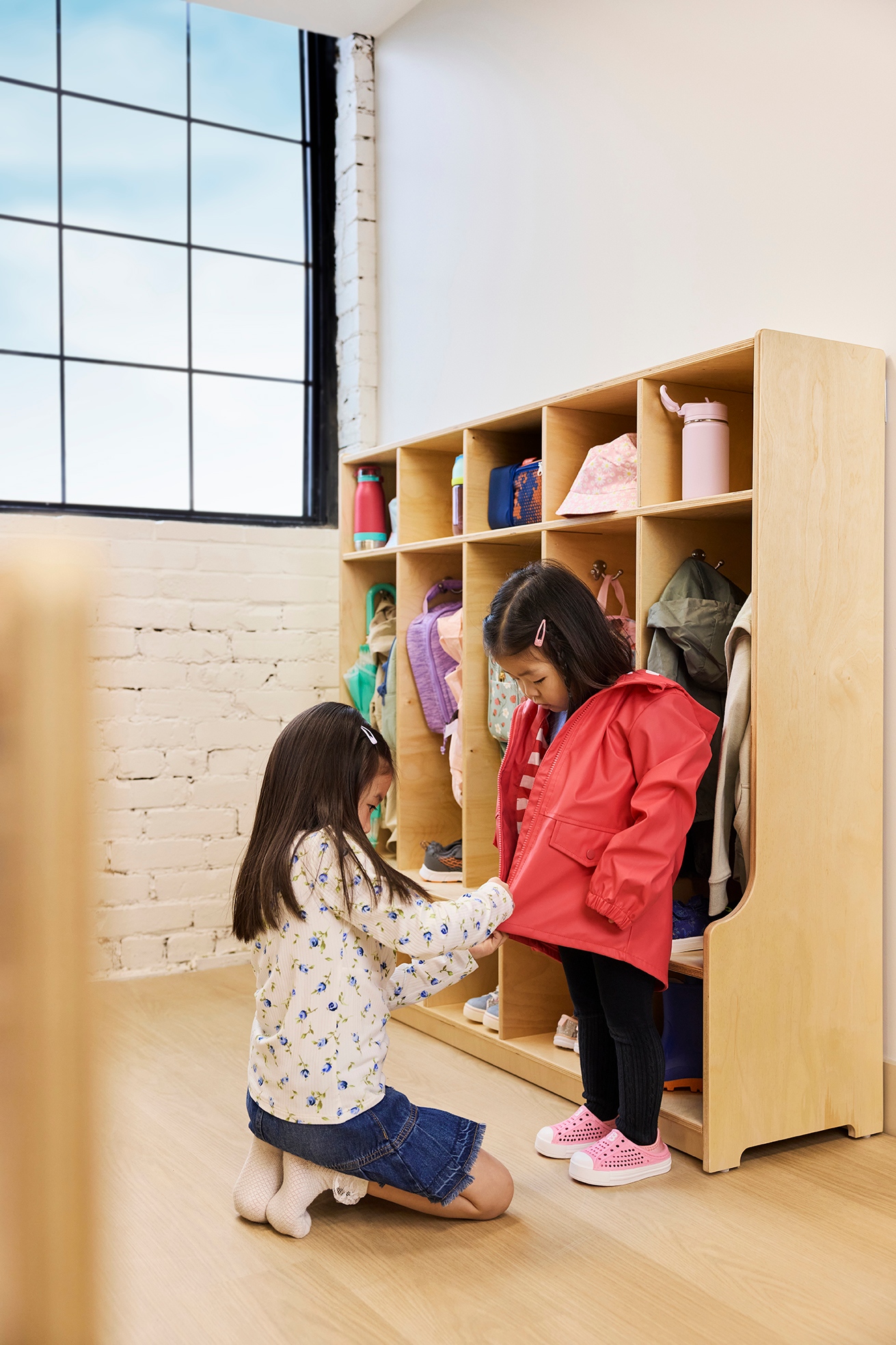 Two girls standing in front of a shelf with childrens belongings. A younger girl is helping an older girl zip up her jacket